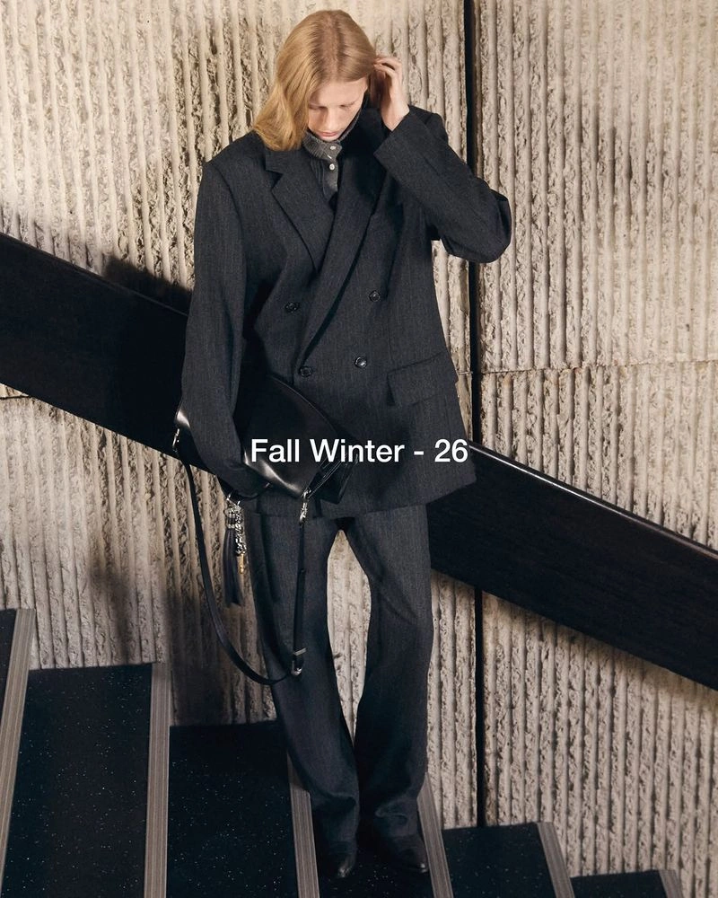 Model wearing an oversized charcoal grey double-breasted suit and holding a black leather bag on a staircase.