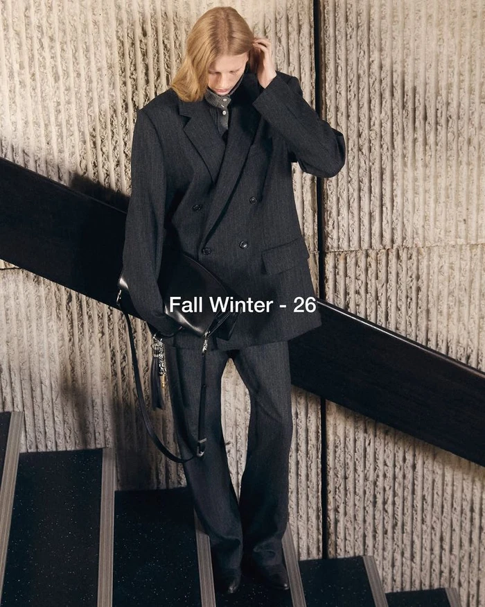 Model wearing an oversized charcoal grey double-breasted suit and holding a black leather bag on a staircase.