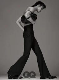 Model Taemin Park walking in profile wearing black high-waisted flared trousers and carrying a black bag against a grey seamless backdrop.