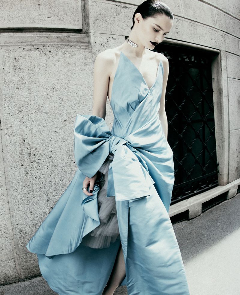 Model in a draped ice blue satin gown with an oversized bow walking outdoors alongside a stone building.