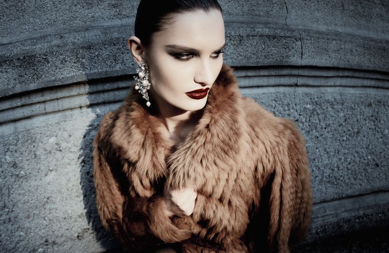 Model wearing a brown fur coat and ornate crystal drop earring posing against a textured stone wall in direct sunlight.