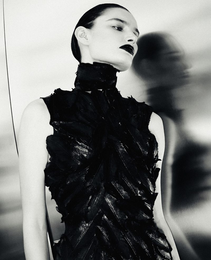 Black and white fashion portrait of a woman in a textured feather high-neck dress against a reflective metallic wall.