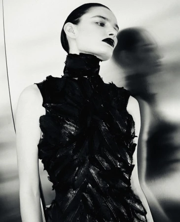 Black and white fashion portrait of a woman in a textured feather high-neck dress against a reflective metallic wall.