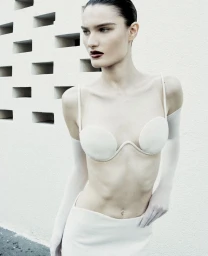 Model wearing a sculptural white bralette, matching skirt, and sheer white arm sleeves against a perforated wall.