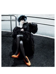 Fashion model wearing a voluminous black feathered coat, black headpiece, and bright orange fuzzy shoes seated on asphalt.