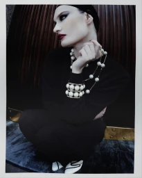 Model in a black outfit and oversized pearl jewelry crouching against a dark fluted wall for Vogue Czechoslovakia.