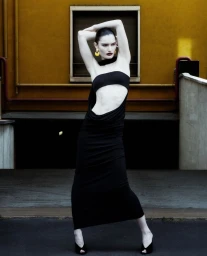 Model posing in a black cut-out dress and yellow earrings with arms raised against an ochre wall and dark architectural entrance.