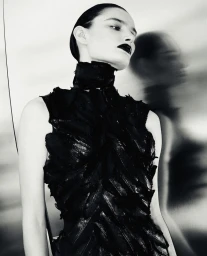 Black and white fashion portrait of a woman in a textured feather high-neck dress against a reflective metallic wall.