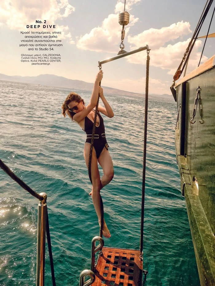 Model wearing a black one-piece swimsuit and sunglasses holds onto a rope swing hanging from a boat over clear turquoise water.