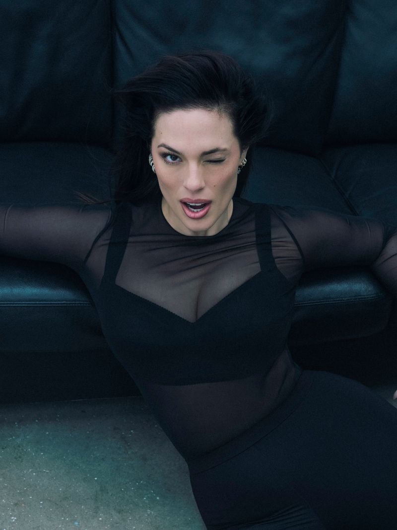 Ashley Graham winking and posing in a black sheer top against a dark leather couch for Elle Spain