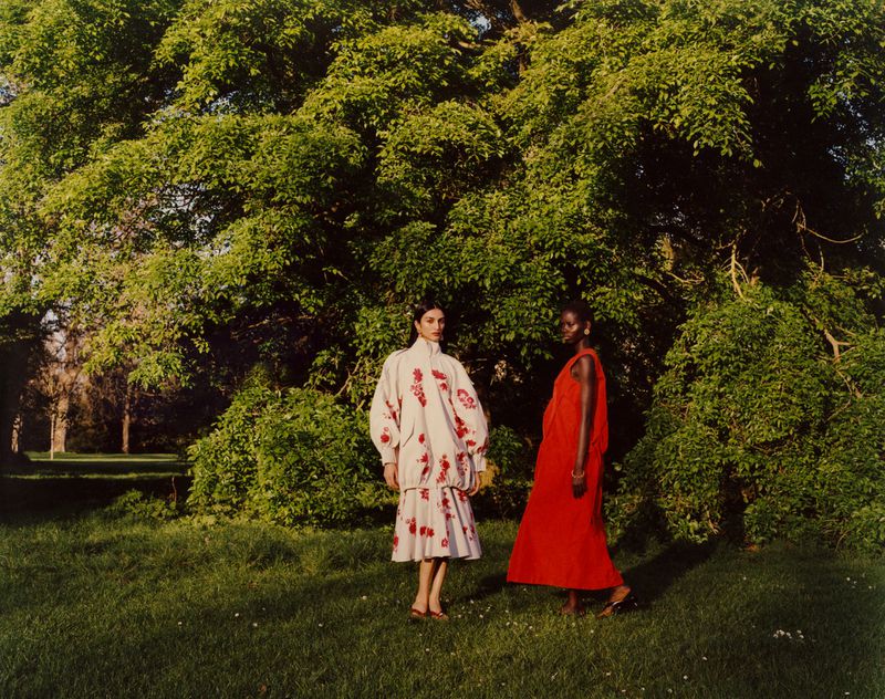 Two female models posing on a grassy lawn in front of a large leafy tree for British Vogue, wearing white patterned and red dresses.