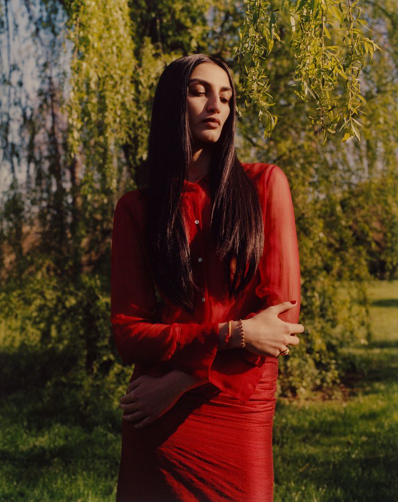 Model Avanti Nagrath wearing a sheer red button-up shirt and red skirt outdoors in warm sunlight for British Vogue.