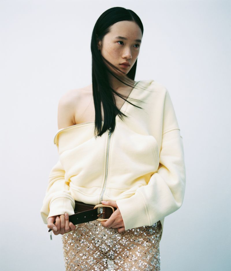 Model with long dark hair wearing an off-the-shoulder cream top and sequin skirt against a neutral background.