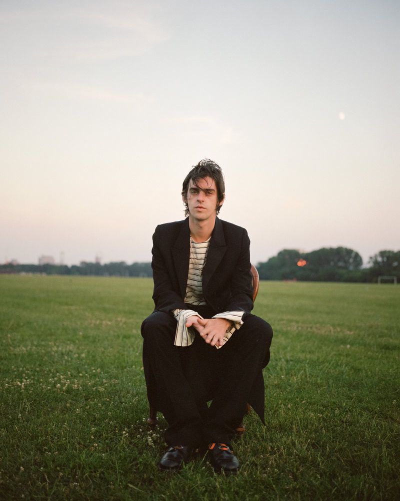 Actor Earl Cave in a black blazer and striped shirt sitting on a chair in an open grassy field at twilight.