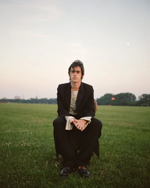Actor Earl Cave in a black blazer and striped shirt sitting on a chair in an open grassy field at twilight.