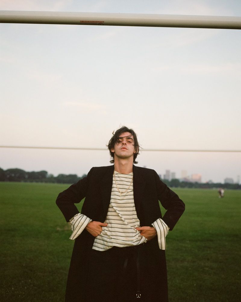 Actor Earl Cave standing in a grassy field wearing an oversized black coat over a striped long-sleeve top.