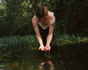 Earl Cave wearing a white tank top and cupping water in a river surrounded by lush foliage.