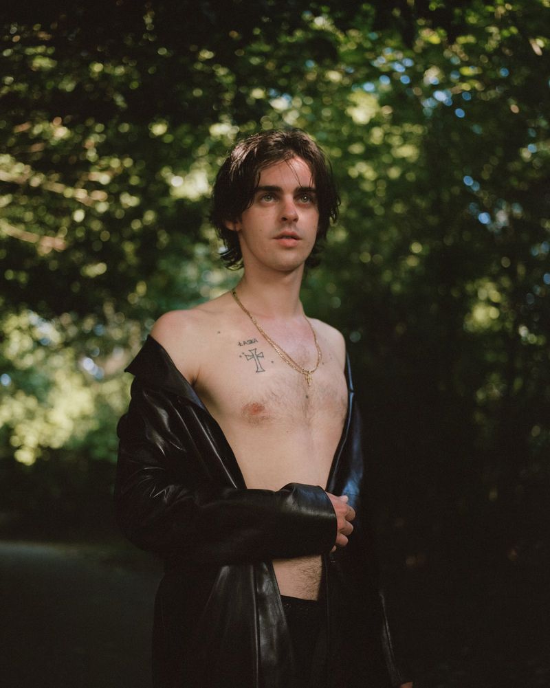 Actor Earl Cave posing shirtless in an open black leather jacket and gold chain necklace outdoors against lush green foliage.