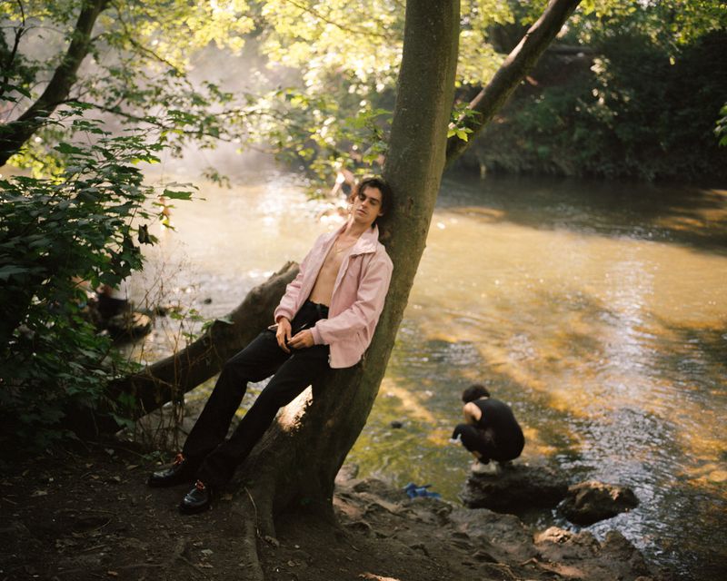 Earl Cave leans against a tree trunk in an open pale pink jacket by a sunlit riverbank.