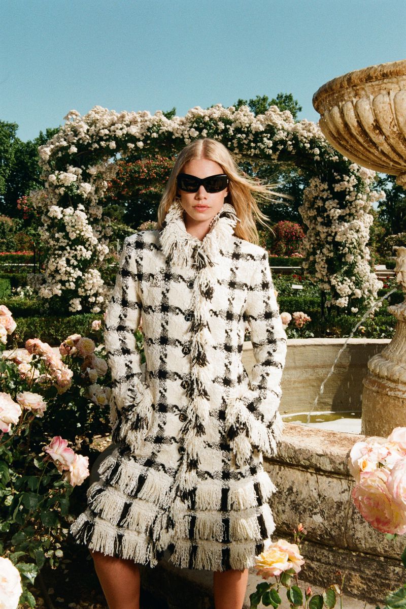 A blonde model wearing a fringed black and white plaid tweed coat and dark sunglasses stands near a stone fountain in a rose garden.