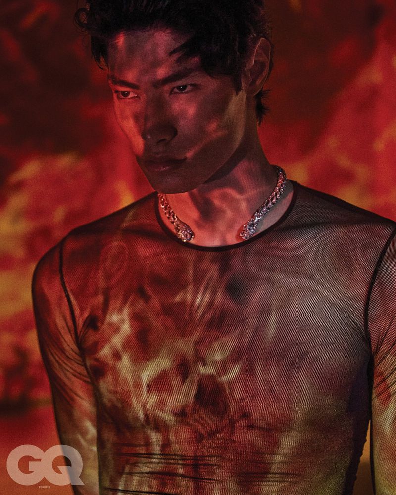 Male model wearing a sheer mesh top and silver dragon necklace illuminated by red flame projections for GQ Turkey.