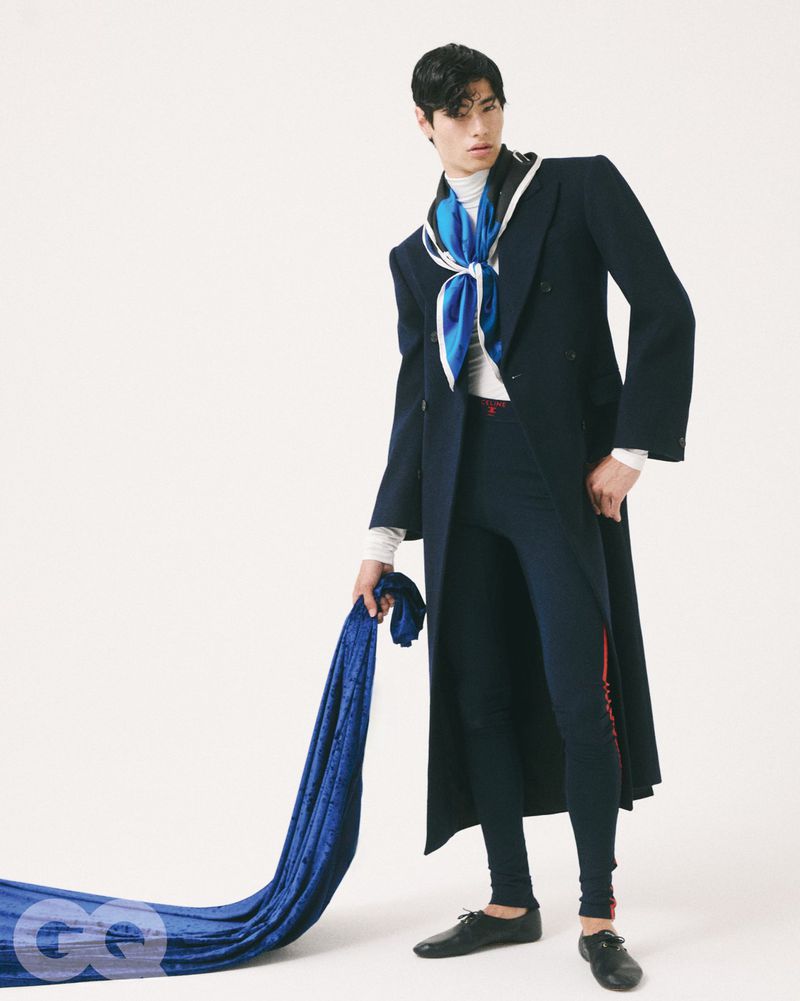 Model Brando Erba posing in a long dark navy coat and blue silk neck scarf holding draped blue velvet against a white studio backdrop.