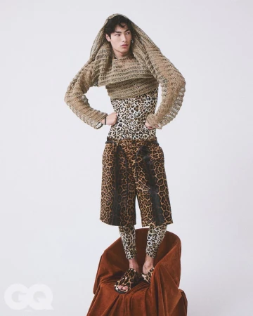 Model standing on a chair in a hooded open-knit sweater and leopard-print layered outfit against a white background.