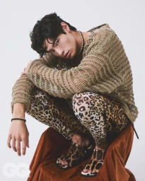 Model crouching on a velvet draped chair wearing a loose open knit sweater and leopard print pants against a white background.