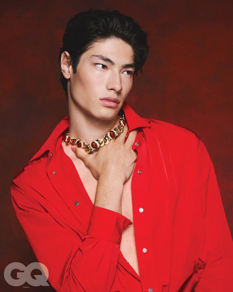Model Brando Erba posing in an unbuttoned bright red shirt and chunky gold stone necklace against a dark red backdrop.