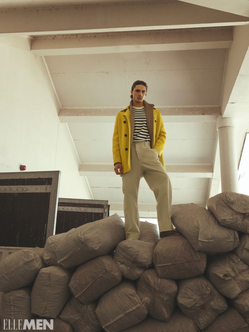 Kamal Sad stands atop a large stack of burlap sacks wearing a bright yellow jacket, striped t-shirt, and beige trousers.