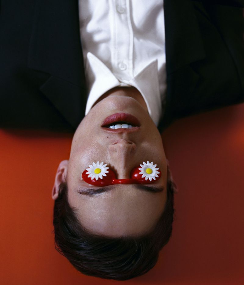 Close-up of model Paulina Kube inverted wearing novelty red glasses with daisies, a black suit jacket, and white collared shirt.