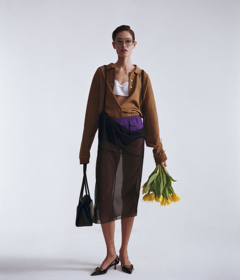 Model Paulina Kube standing in a brown shirt, sheer black skirt, and glasses holding yellow flowers against a white seamless background.