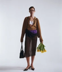 Model Paulina Kube standing in a brown shirt, sheer black skirt, and glasses holding yellow flowers against a white seamless background.