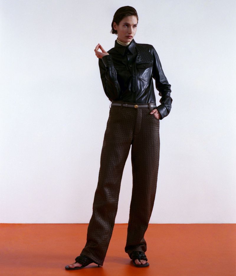 Model Paulina Kube standing in a black leather shirt and brown textured trousers on an orange floor against a white studio backdrop.