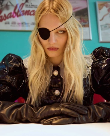 Blonde woman wearing a black eye patch and dark glossy dress with white lace collar against a vibrant turquoise background.