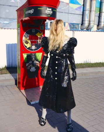 Blonde woman in shiny black coat and long gloves standing outdoors beside a red punching bag machine for Vogue Portugal.