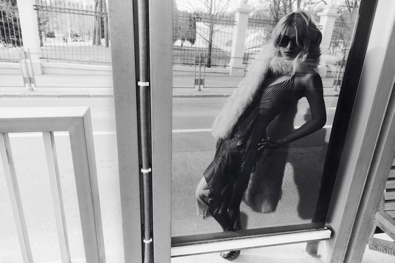 Black and white portrait of a woman wearing sunglasses and a furry scarf, leaning against a glass bus shelter wall.
