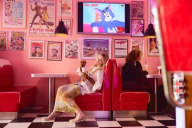 Woman in an oversized white shirt and long gloves reclining in a red vinyl booth inside a retro pink diner.
