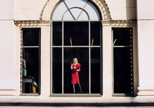 Woman wearing a vibrant red dress and black gloves stands behind a tall arched window set in a light stone building facade.