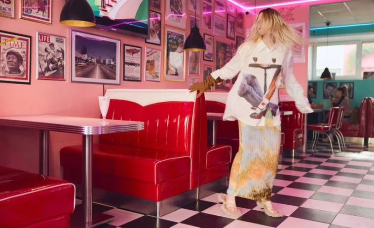Model with spinning blonde hair dancing between glossy red vinyl booths inside a retro diner with a checkered tile floor.