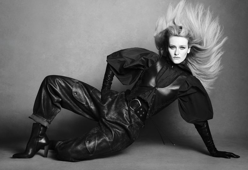 Actor Sarah Pidgeon poses reclining on the floor in a voluminous black leather outfit with windswept hair against a grey backdrop.