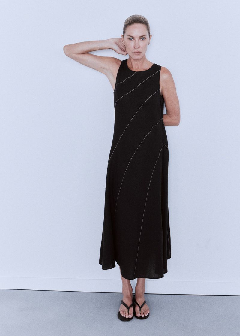 Model Erin Wasson wearing a sleeveless black midi dress with diagonal topstitching and black sandals against a white background.