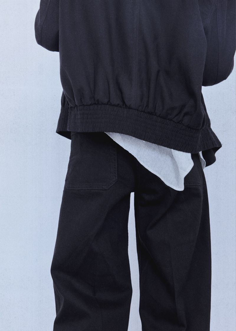 Back view of a model wearing a black bomber jacket and matching black trousers with a white top hem against a seamless backdrop.