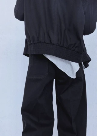 Back view of a model wearing a black bomber jacket and matching black trousers with a white top hem against a seamless backdrop.