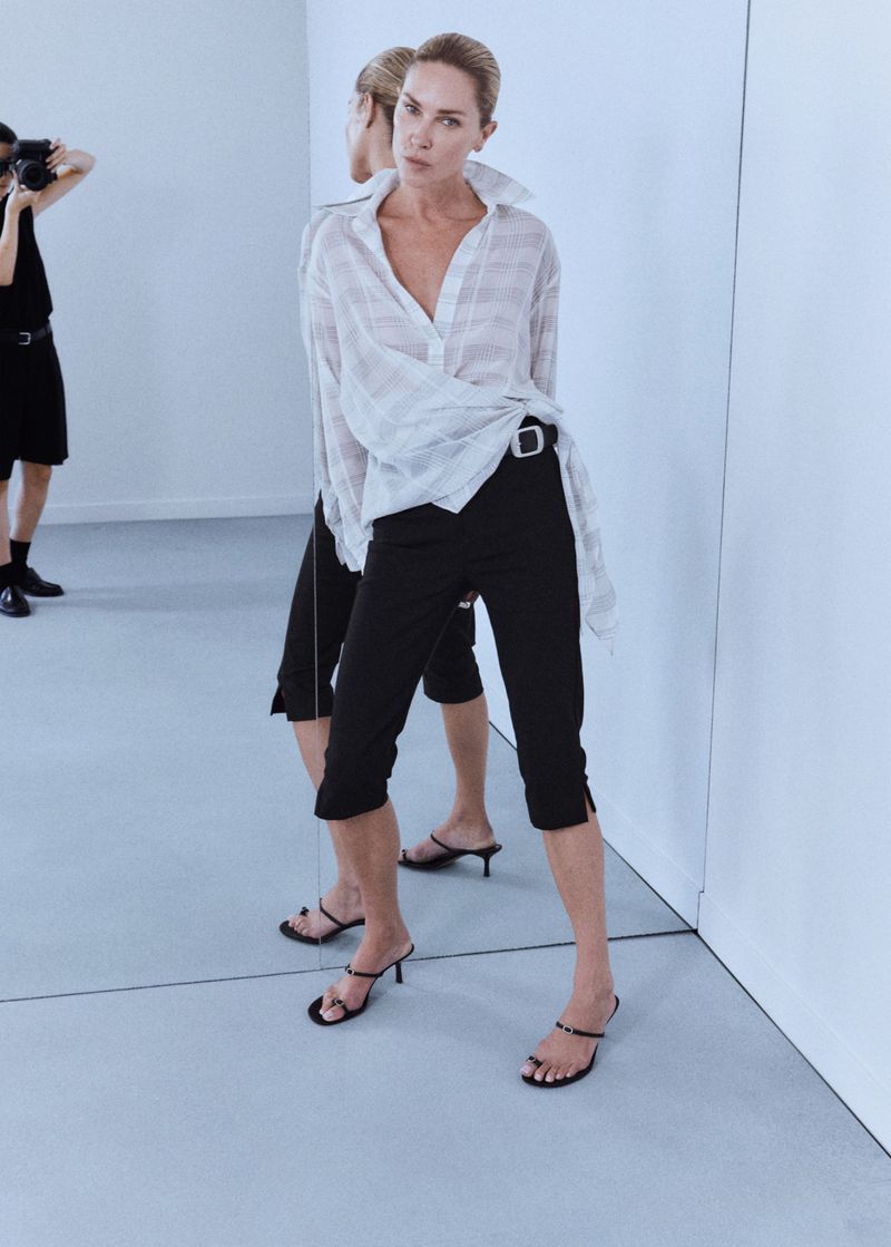 Model Erin Wasson posing before a mirror in a sheer striped white shirt, black capri pants, and strappy kitten heels.