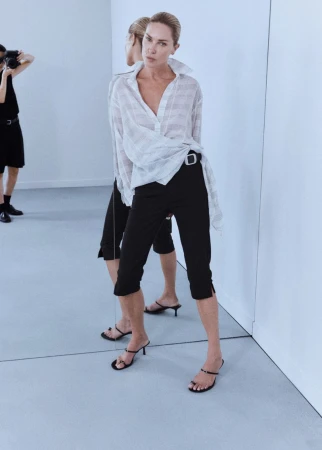 Model Erin Wasson posing before a mirror in a sheer striped white shirt, black capri pants, and strappy kitten heels.