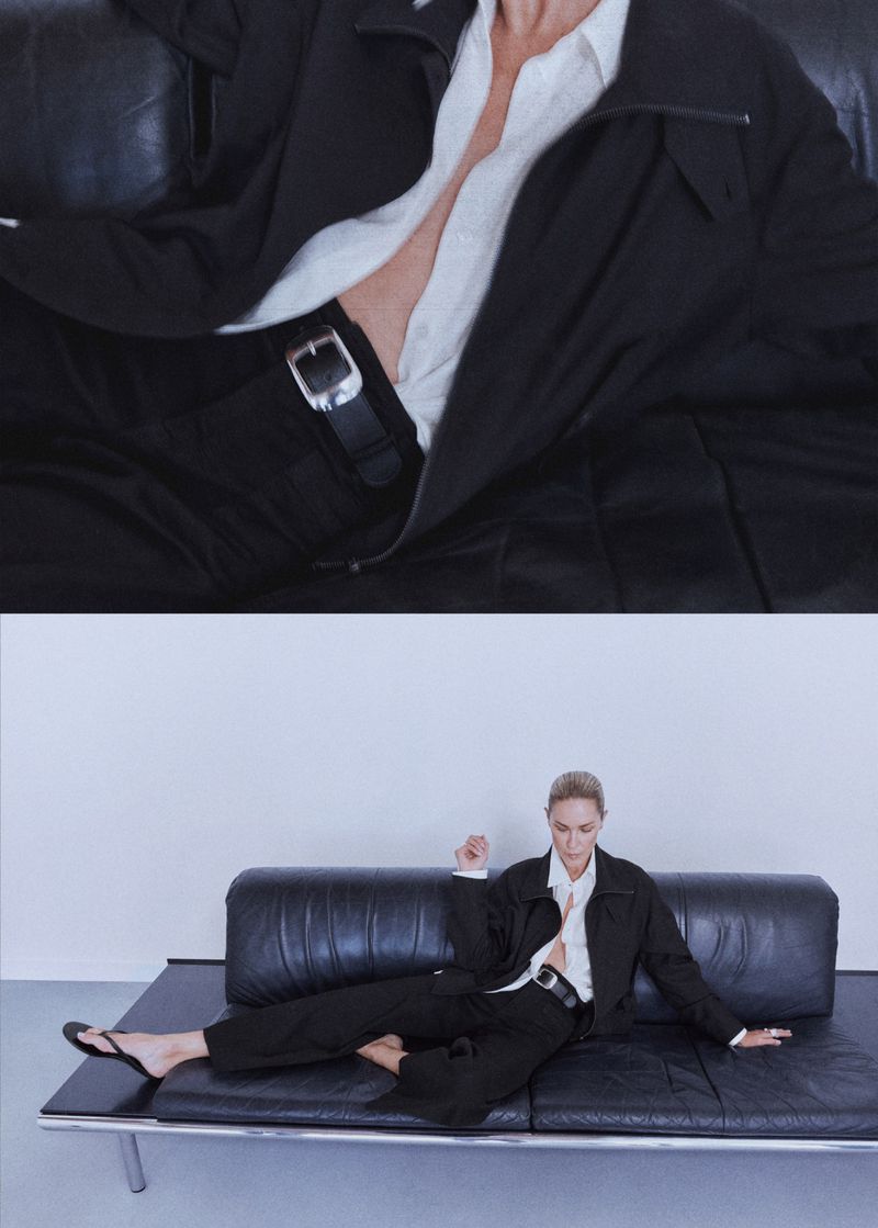 Model Erin Wasson wearing a black jacket, unbuttoned white shirt, and black trousers while lounging on a black leather daybed.