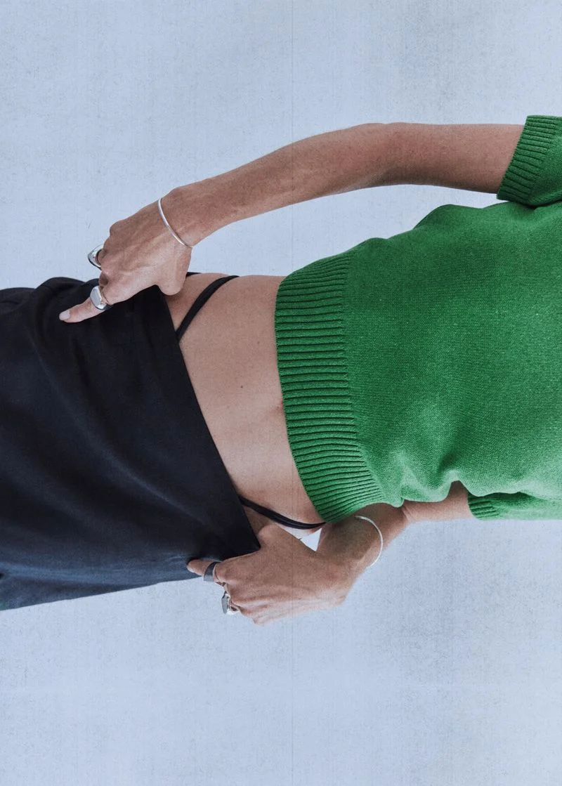 Erin Wasson wearing a bright green short-sleeve knit sweater and black trousers against a pale background.