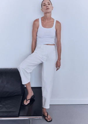 Model Erin Wasson wearing a white tank top, white trousers, and black flip flop sandals with one foot on a black leather bench.
