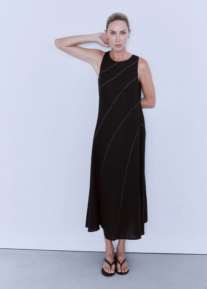 Model Erin Wasson wearing a sleeveless black midi dress with diagonal topstitching and black sandals against a white background.